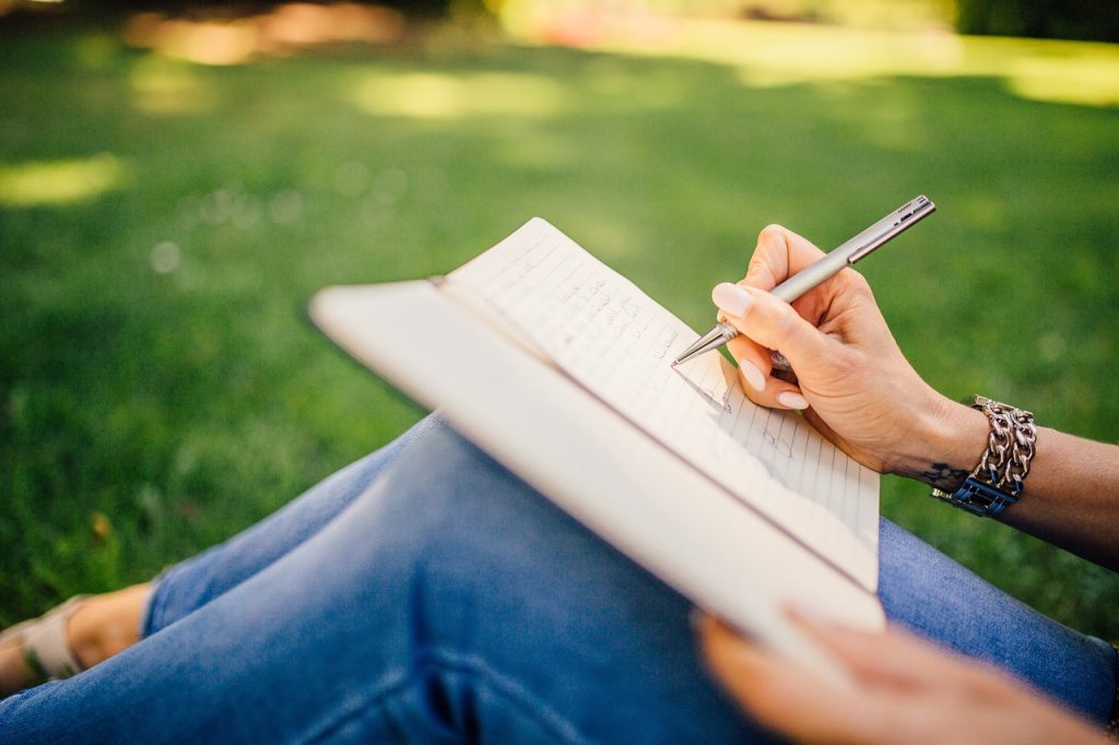Gros plan sur la main d'une femme assise dans l'herbe en train d'écrire dans un carnet.